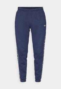 Granatowe spodnie dresowe z elastycznym pasem i ściągaczami, z białymi bocznymi paskami z wzorem logo oraz małym zielonym logo marki na nogawce.