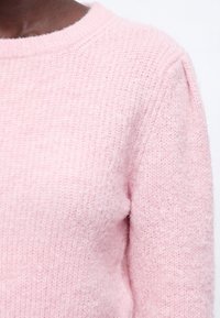 Pull en maille rose doux porté sur la partie supérieure du corps montrant un tissu texturé et un col rond sur fond blanc.