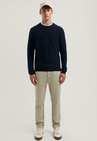 Marineblauer Strickpullover, beigefarbene Hose und weiße Sneaker. Das Model trägt eine helle Mütze. Der Pullover hat einen runden Halsausschnitt und gerippte Bündchen.