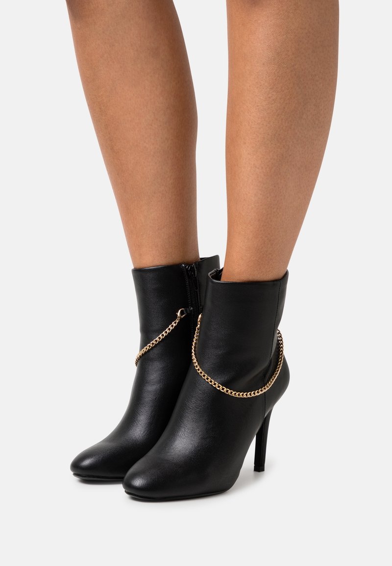 Bottes basses en cuir noir avec détail chaîne dorée et bouts pointus, portées sur jambes nues sur fond blanc.