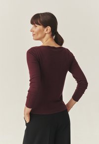 Burgunderfarbener taillierter Langarmoberteil mit rundem Halsausschnitt und gerippten Bündchen, kombiniert mit schwarzen, hochtaillierten Hosen. Weicher Stoff, nahtloses Design.