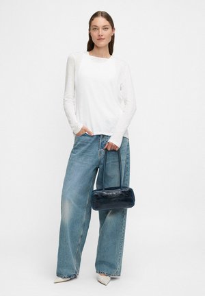 Una donna che indossa un top bianco a maniche lunghe e jeans a gamba larga blu, tiene in mano una piccola borsa blu scuro, in piedi contro uno sfondo bianco uniforme.