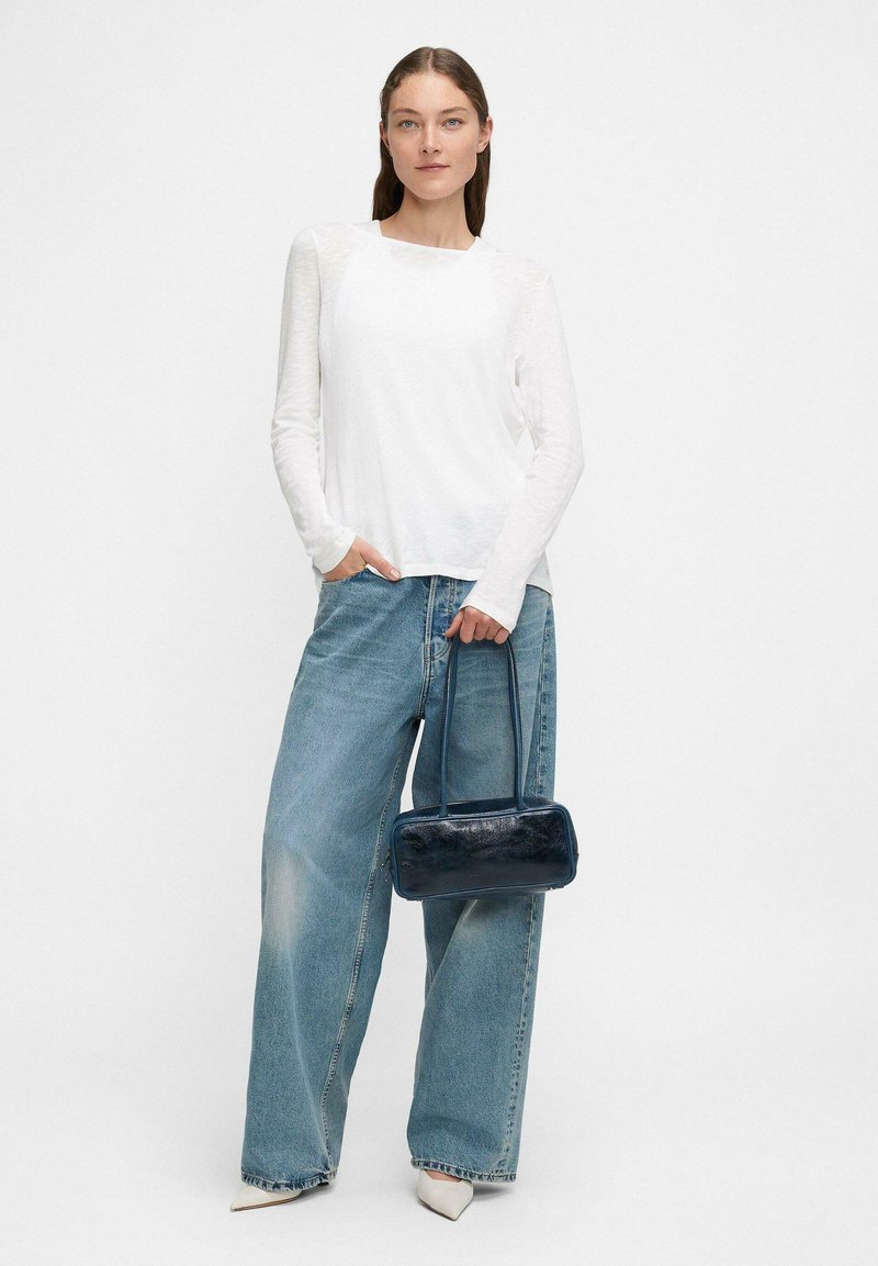Femme portant un haut blanc à manches longues et un pantalon en jean bleu ample, tenant un petit sac à main bleu marine, se tenant contre un fond blanc uni.