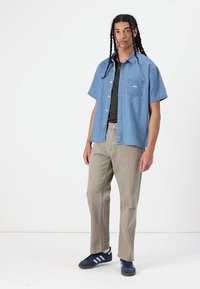 Short-sleeve denim shirt in light blue, with a front pocket and white logo, paired with light beige pants and dark blue sneakers.