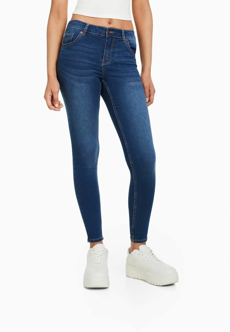 Jeans de denim azul ajustados con una cintura de media altura, que presentan un estilo tradicional de cinco bolsillos y detalles de costura sutiles. Llevados con zapatillas blancas de plataforma.