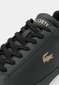 Črne usnjene superge s črnimi vezalkami, ki na strani vsebujejo zlat napis "Lacoste" in emblem krokodila. Gladka tekstura in minimalističen dizajn.