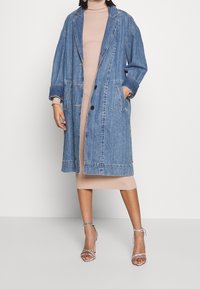 Veste en denim bleu clair au design long et ample, avec de grands boutons et des poches latérales ; portée par-dessus une robe beige ajustée.