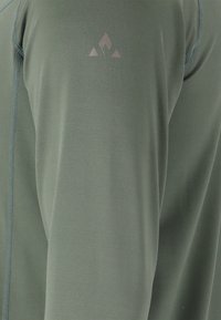 Nahaufnahme von olivgrünem Sportstoff mit sichtbaren Nähten und einem kleinen reflektierenden geometrischen Logo am Ärmel.