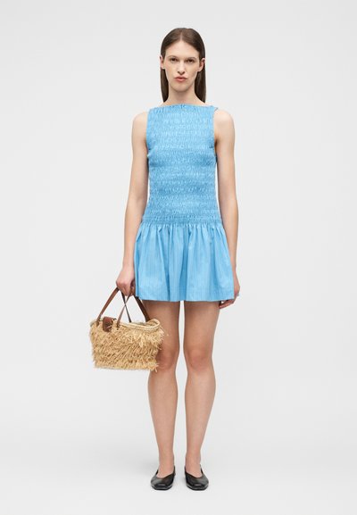 Γυναίκα με μπλε φόρεμα χωρίς μανίκια, με σφηκοφωλιά, κρατώντας ένα πλεκτό καλαθάκι από ψάθα με δερμάτινα χερούλια, φοράει μαύρα φλατ παπούτσια, στέκεται σε ένα απλό φόντο.