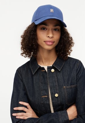 Persoană tânără cu părul creț, purtând o șapcă de baseball albastră și o geacă din denim închis la culoare, cu brațele încrucișate, pe un fundal simplu.