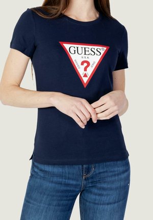 Kobieta w granatowej koszulce Guess z czerwono-białym trójkątnym logo i niebieskich dżinsach, ręce luźno splecione na wysokości talii.