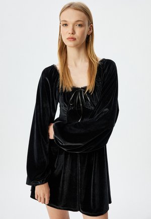Black velvet dress with long, puffed sleeves, square neckline, and a bow detail at the front. Features a flowy silhouette and lace accents.