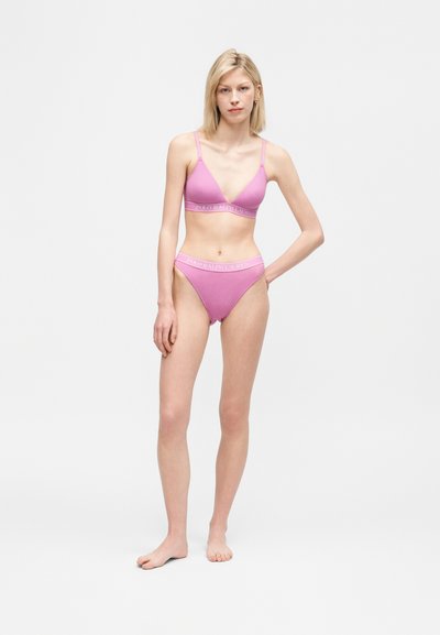 Νεαρή γυναίκα όρθια ξυπόλητη, φοράει ροζ σετ εσωρούχων Polo Ralph Lauren με ελαστικές ταινίες με το λογότυπο, σε λευκό φόντο.
