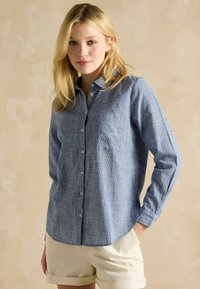 Lichtblauw geruit overhemdd met lange mouwen, zachte stof en één zak; gecombineerd met crèmekleurige shorts.