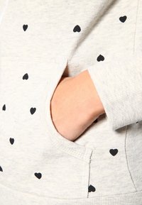 Sudadera con capucha gris claro hecha de tela suave y texturizada con pequeños patrones de corazones negros. Incluye un bolsillo delantero y puños acanalados.