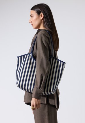 Ruskeaan asuun pukeutunut nainen kantaa suurta tummansinivalkoraidallista olkalaukkua ja katsoo vasemmalle yksiväristä taustaa vasten.