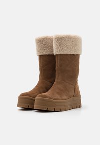 Bruna mockasiner i ankelhöjd med en fyllig beige fårluddskant, featuring en tjock plattformssula med ribbad detalj.