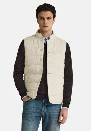 Ung mand iført en beige quiltet vest med knapper over en sort sweater og blå jeans, stående med den ene hånd i lommen mod en hvid baggrund.