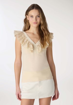 Młoda kobieta z długimi falującymi włosami, ubrana w beżowy, falbaniasty top na ramiączkach z dekoltem w serek i białą mini spódniczkę, stojąca na jednolitym tle.