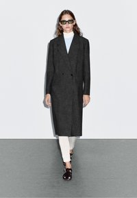 Dvojřadový tmavě šedý kabát s klopami, nošený přes světle modrý rolák a krémové kalhoty, doplněný zelenými slunečními brýlemi a černými sandály.