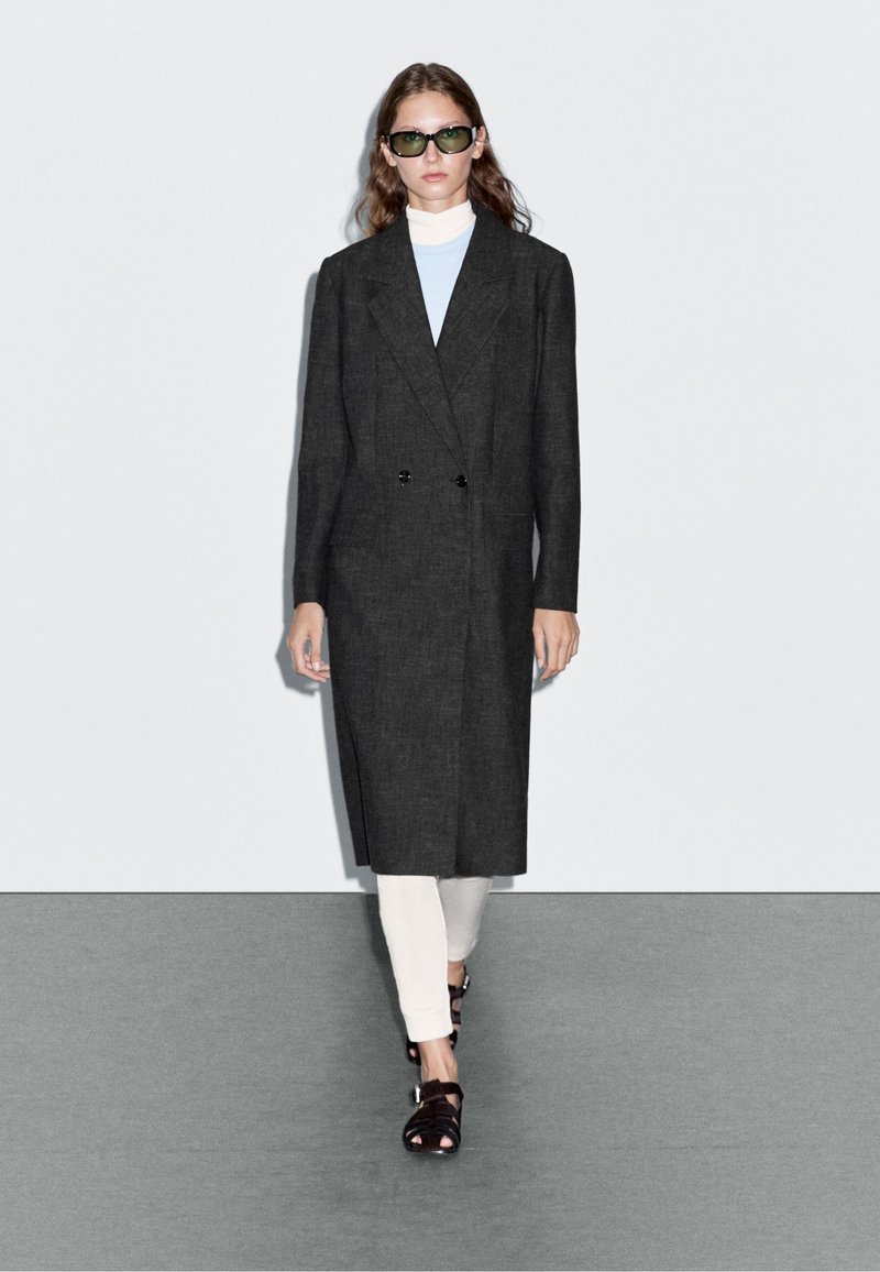 Dvojřadový tmavě šedý kabát s klopami, nošený přes světle modrý rolák a krémové kalhoty, doplněný zelenými slunečními brýlemi a černými sandály.