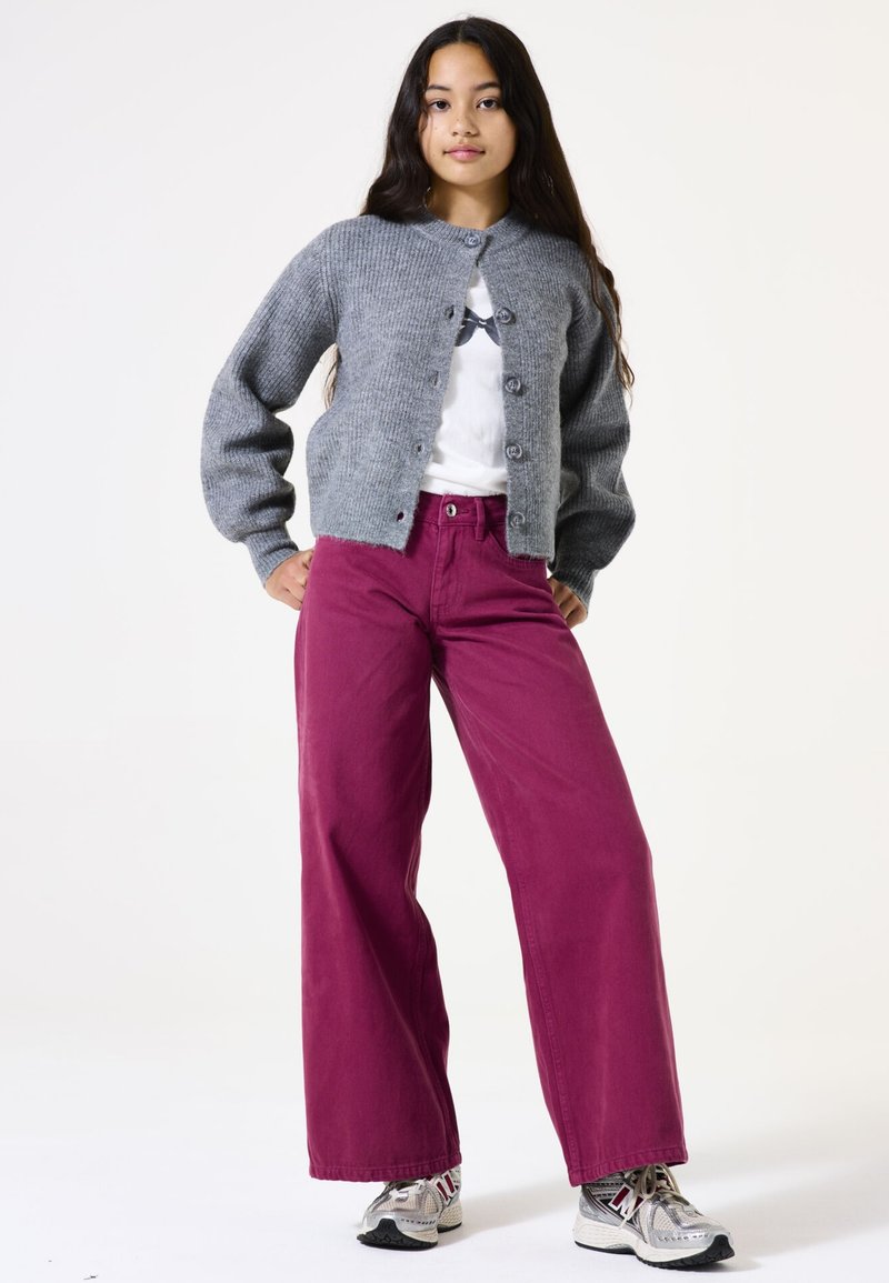 Grauer gestrickter Cardigan mit rundem Ausschnitt und Knöpfen, kombiniert mit weit geschnittenen fuchsia-farbenen Hosen und grauen Sneakers mit Details. Einfacher Hintergrund.