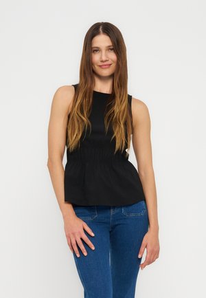 Kobieta o długich brązowych włosach, ubrana w czarny top bez rękawów i niebieskie dżinsy, stoi na tle jasnego, jednolitego tła, uśmiechając się.