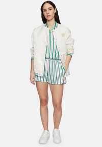 Chaqueta bomber de satén blanca con cuello a rayas verdes y blancas, combinada con una camiseta y pantalones cortos a rayas verdes y moradas a juego. Zapatillas blancas.