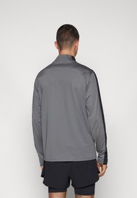 Langarm-Sportoberteil aus grauem Stoff mit schwarzen Seitenelementen, mit hohem Kragen und glatter Textur, getragen mit dunklen Shorts.