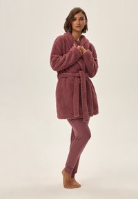 Puszysty, różowy szlafrok o teksturowanej powierzchni i pasku, noszony na dopasowanych spodniach. Gładki materiał podkreśla luźny krój.