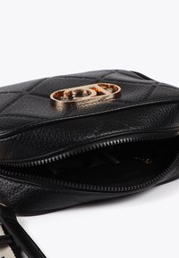 Svart vadderad axelväska med ett guld-logoknäpp, delvis öppen för att avslöja ett mörkt inre och dragkedje-detaljer.