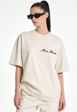 Woman with black ponytail wearing black rectangular sunglasses and an oversized beige T-shirt with "Mira Paris" text on front.