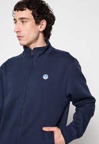 Giacca in pile blu navy con cerniera, colletto alto, polsini a coste e logo circolare sul lato sinistro del petto. Tessuto morbido e vestibilità rilassata.