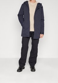 Mies, joka pukeutunut tummansiniseen hupulliseen takkiin, beigeen teksturoituun neulepaitaan, mustiin cargo-housuihin ja mustiin nyörikenkiin, seisoo valkoista taustaa vasten.