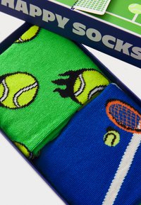Gröna strumpor med gula tennisboll och flamestilsmönster, parat med blå strumpor med design av tennisracket och boll. Presenterades i en låda.