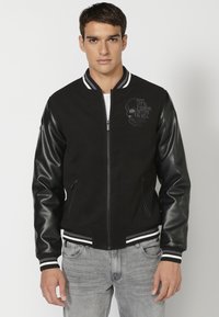 Black bomber jacket with leather sleeves, ribbed cuffs and hem, featuring a skull graphic and text on the chest. Zipper front, side pockets.