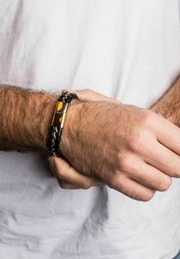 Fekete fonott bőrkarkötő arany tónusú záróelemmel és kiegészítőkkel. Fényes felülettel rendelkezik, és téglalap alakú középső része van.