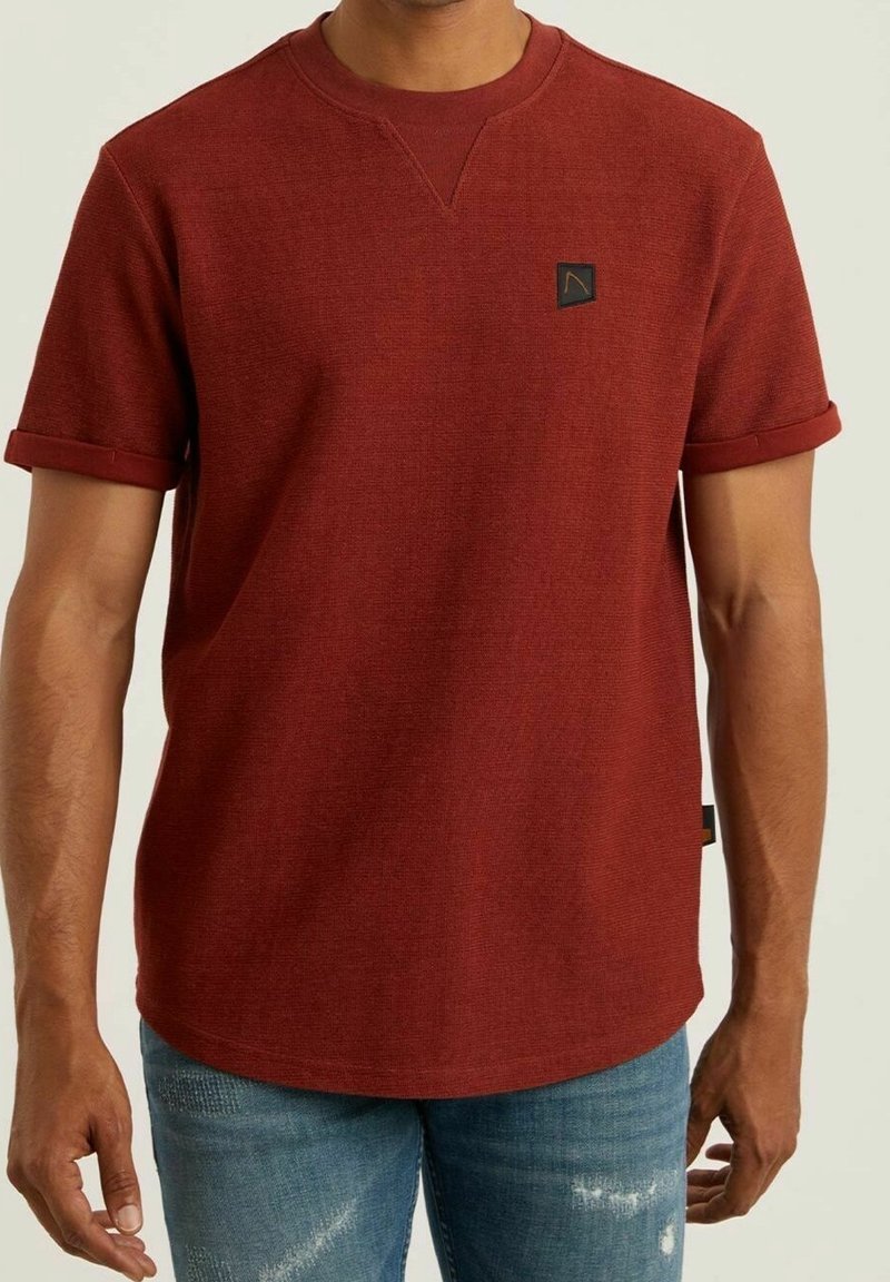 Rotes strukturiertes T-Shirt mit kurzen Ärmeln, rundem Halsausschnitt und umgeschlagenen Bündchen, das ein kleines schwarzes Logopatch auf der Brust zeigt.