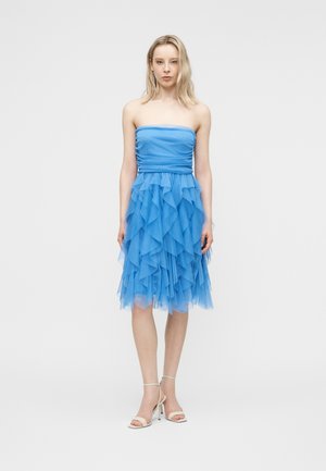 Vaaleahiuksinen nainen seisoo olkaimettomassa, polvipituisessa sinisessä mekossa, jossa on kerroksittaiset röyhelöt, ja valkoisissa korkokengissä yksiväristä taustaa vasten.