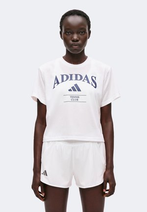 Kobieta w białej koszulce Adidas Tennis Club i dopasowanych białych krótkich spodenkach sportowych stoi na jednolitym jasnym tle.