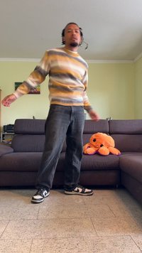 Jeans grigi, sneakers Nike in bianco e nero e un maglione a righe con giallo, grigio e bianco. Un polpo di peluche arancione è seduto su un divano marrone.