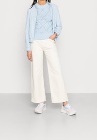 Lichtblauwe bomberjack, blauw truitje met diamantpatroon, witte broek met brede pijpen en pastelkleurige sneakers met grijze en gele accenten.