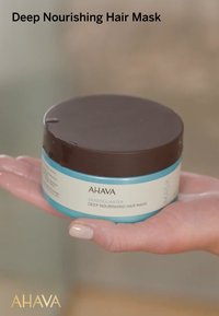 Brązowy i turkusowy okrągły pojemnik z białą etykietą; na etykiecie napisane "AHAVA Głęboko Odżywiająca Maska do Włosów" z wymienionymi składnikami.