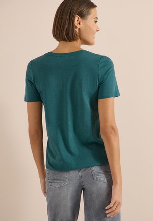 Femme aux cheveux bruns courts portant un t-shirt à manches courtes turquoise et un jean gris, vue de dos sur un fond beige uni.