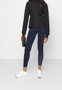 Kvinna iklädd svart zip-up jacka, marinblå leggings, vita sneakers, hållande svart vattenflaska, stående på en ljusgrå golv- och väggbakgrund.