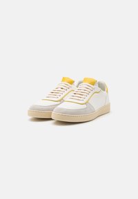Baskets en cuir blanches et gris clair avec des accents jaunes, design à lacets, bout perforé et semelle texturée. Style minimaliste.