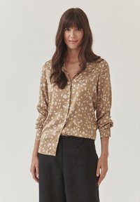 Bruine button-up shirt met witte abstracte vlekken, lange mouwen opgerold bij de manchetten, gecombineerd met zwarte wijde broekspijpen. Zachte textuur.