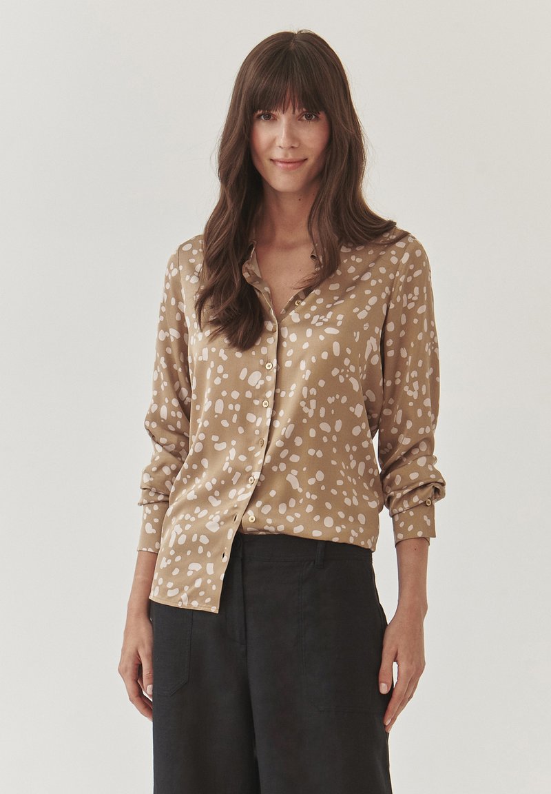 Bruine button-up shirt met witte abstracte vlekken, lange mouwen opgerold bij de manchetten, gecombineerd met zwarte wijde broekspijpen. Zachte textuur.