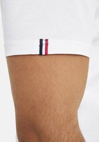 Weißes Baumwoll-T-Shirt mit einer Nahaufnahme des Ärmels, der rot-weiß-blaue gestickte Streifen nahe dem Saum zeigt. Glatte Textur.