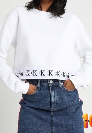 Personne portant un sweat-shirt court blanc avec le logo noir "cK" répété sur l'ourlet et une jupe en denim bleue taille haute avec des bandes rouges sur les côtés.